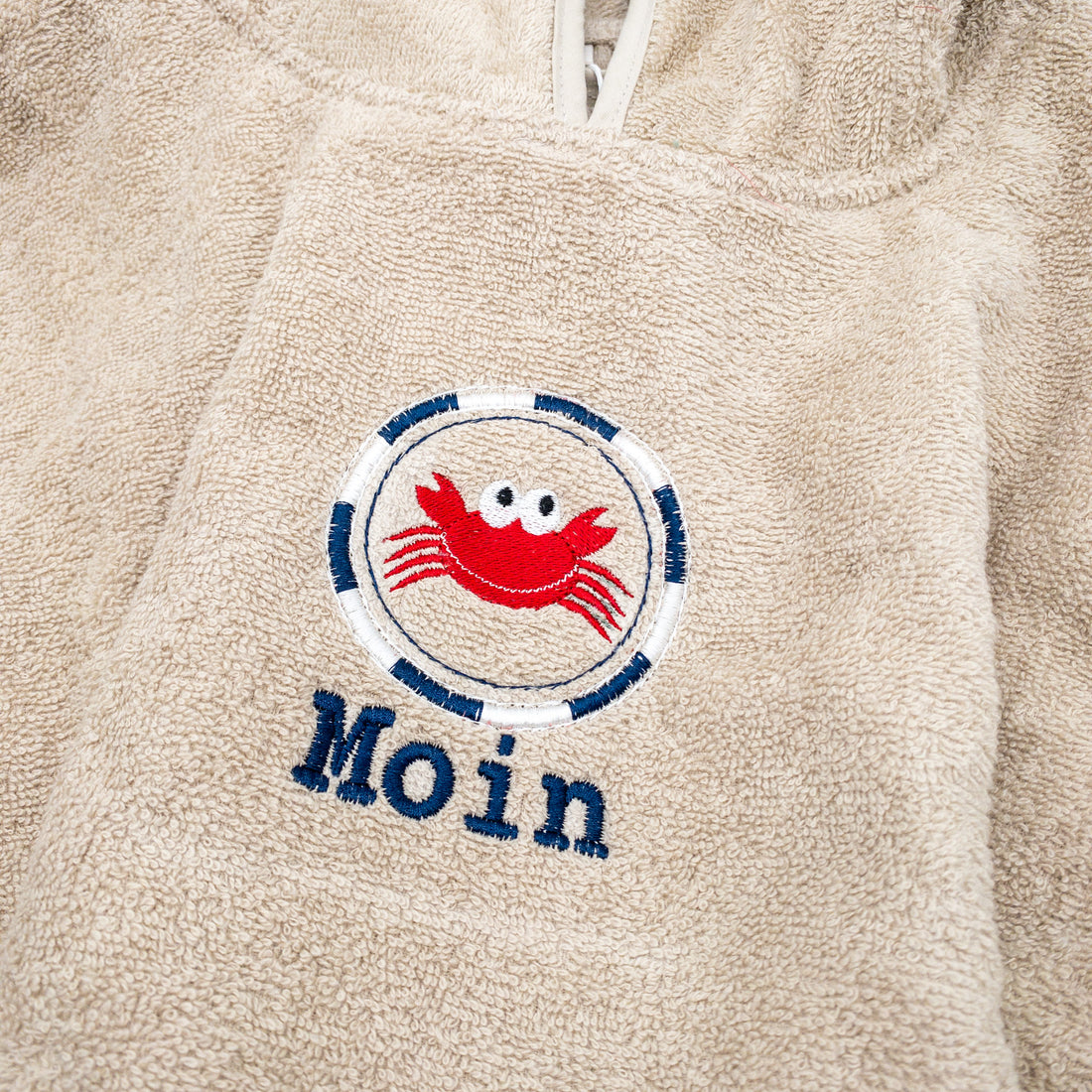 Badeponcho von Krabbelhelden, beige mit Namen und Krebs bestickt-Motiv in Nahaufnahme.